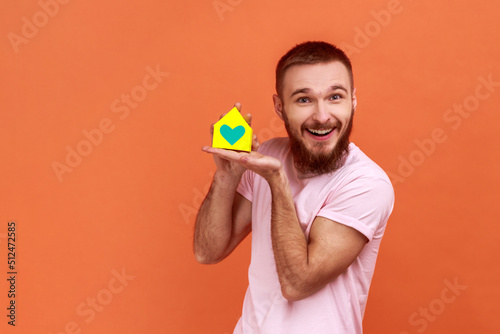 Portrait of bearded man holding in hands and showing paper model of house of his dream, real estate agency advertisement, wearing pink T-shirt. Indoor studio shot isolated on orange background.