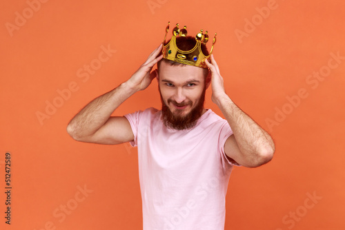 Portrait of handsome bearded man wearing golden crown, looking with positive facial expression, privileged status, wearing pink T-shirt. Indoor studio shot isolated on orange background.