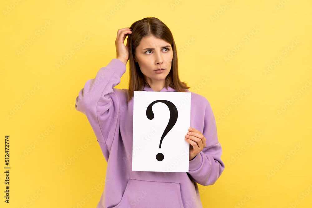 Portrait of pensive thoughtful young woman looking away, holding paper with question mark, thinks about tasks, wearing purple hoodie. Indoor studio shot isolated on yellow background. Portrait of pensive thoughtful young woman looking away, holding paper with question mark, thinks about tasks, wearing purple hoodie. Indoor studio shot isolated on yellow background.