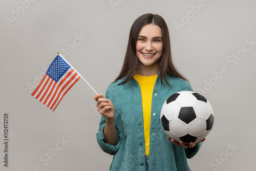 Smiling woman holding flag of united states of america and soccer black and white ball, united soccer league, wearing casual style jacket. Indoor studio shot isolated on gray background. photo