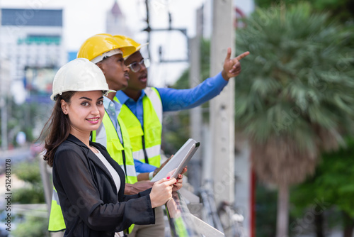 caucasian businesswoman in black suit wear helmet, holding tablet, looking at camera while black engineer colleauges working at construction site outside building in city photo