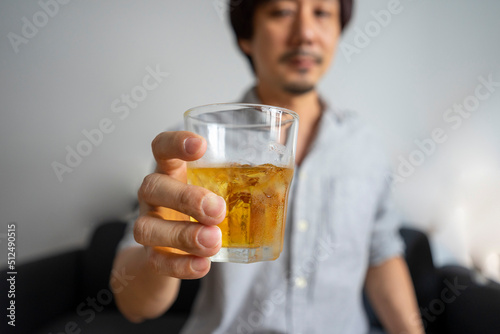Asian man sitting on sofa enjoy a glass of whiskey. Focus on the glass. Alcohol drinking concept.