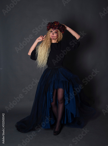 Beautiful Halloween Make-Up Style. Blond curly hair Model Wear Sugar Skull Makeup with Red Roses. Santa Muerte concept. Model on black background in studio with wreath
