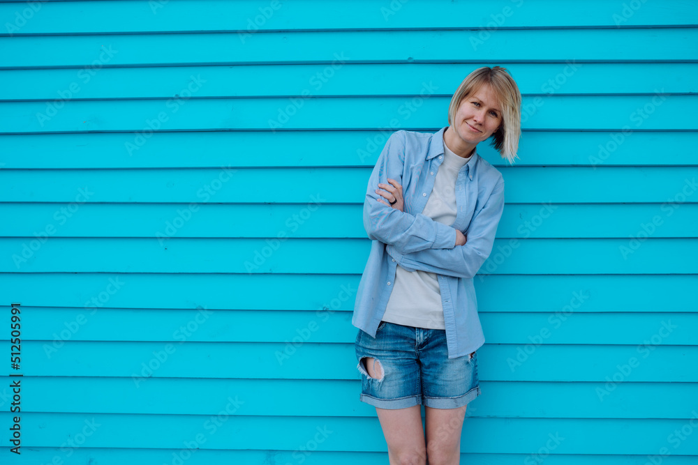 Portrait of beautiful middle-aged smiling woman with short fair hair wearing grey T-shirt, blue shirt, torn denim shorts, leaning against blue wooden wall with crossed arms, bowing head, relaxing. Portrait of beautiful middle-aged smiling woman with short fair hair wearing grey T-shirt, blue shirt, torn denim shorts, leaning against blue wooden wall with crossed arms, bowing head, relaxing.