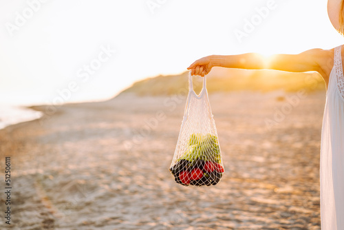 Cropped photo of woman wearing long white summer sundress, standing on sandy beach near water sea ocean on sunny day, stretching hand with string bag full of vegetables. Summer, vacation, holiday.