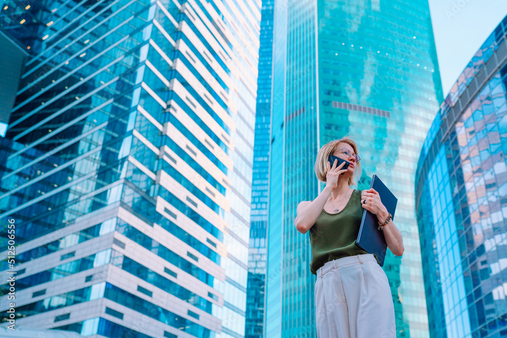 Fototapeta premium Portrait of busy middle-aged woman businesswoman with fair hair wearing green top, white trousers, standing on street in city centre near glass skyscrapers, holding blue laptop, talking on smartphone.