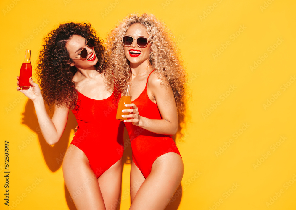 Two beautiful sexy smiling hipster women in red summer swimwear bathing suits.Trendy models with afro curls hairstyle having fun in studio.Hot female isolated on yellow.Drinking lemonade from bottle