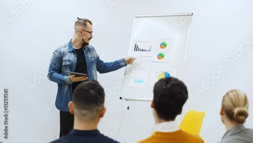 Businessman coach trainer presents report, shows diagrams on flip chart in conference hall. Audience raises hands for answer on presentation in office. Teacher leading master class to audience. photo