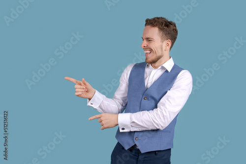 Young business man in white shirt and blue suit vest isolated on blue background studios portrait. Career achievement wealth business concept. Shows a gesture to a copy of the place and smiles