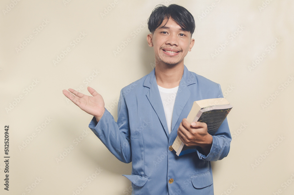 young asian man wearing college suit pointing to empty space while carrying book on isolated background young asian man wearing college suit pointing to empty space while carrying book on isolated background