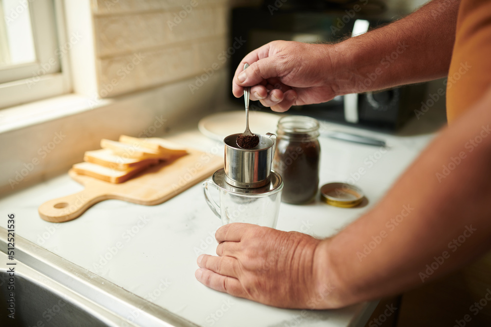 Hands of elderly man adding two spoons of ground coffee in phin filter when making Vietnamese style coffee