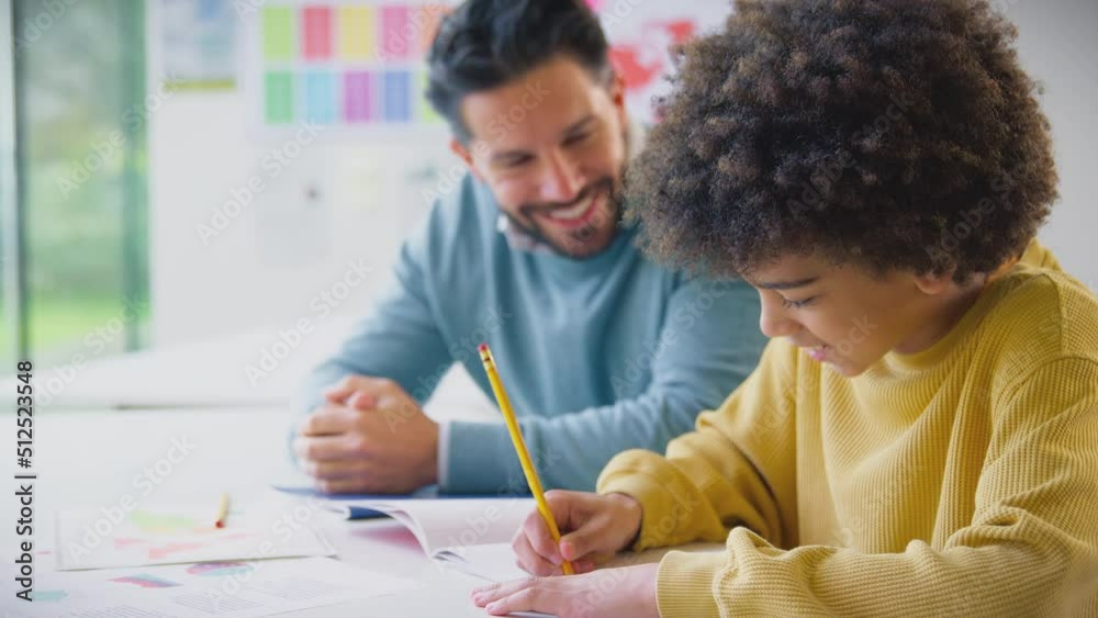 Male teacher sitting at desk with male elementary pupil in classroom as he writes in school exercise book - shot in slow motion Male teacher sitting at desk with male elementary pupil in classroom as he writes in school exercise book - shot in slow motion