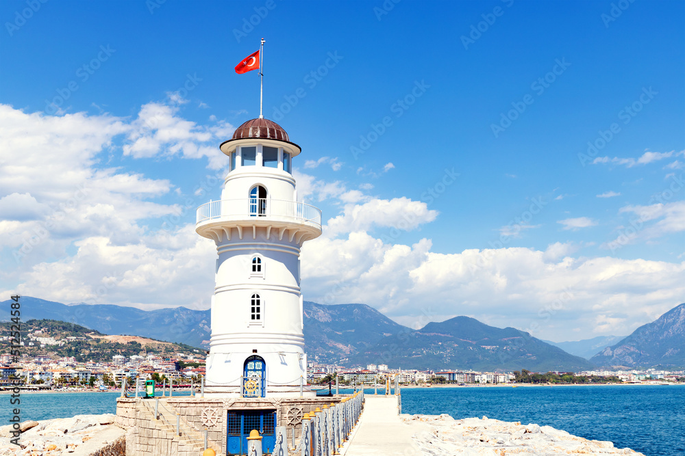 White lighthouse with turkish flag in Alanya, Turkey. Panoramic view of city, sea and harbour under cloudy sky. Selective focus.