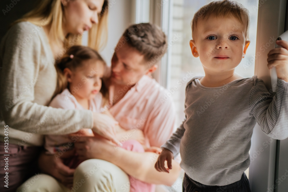 cute caucasian little boy standing on window sill. His parents and sister on background looking out the window. cute caucasian little boy standing on window sill. His parents and sister on background looking out the window.
