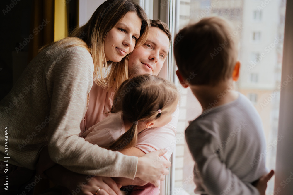 cute caucasian little boy standing on window sill. His parents and sister on background looking out the window. cute caucasian little boy standing on window sill. His parents and sister on background looking out the window.