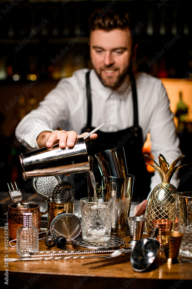 focus on a crystal glass into which male bartender pours a drink from a steel shaker cup