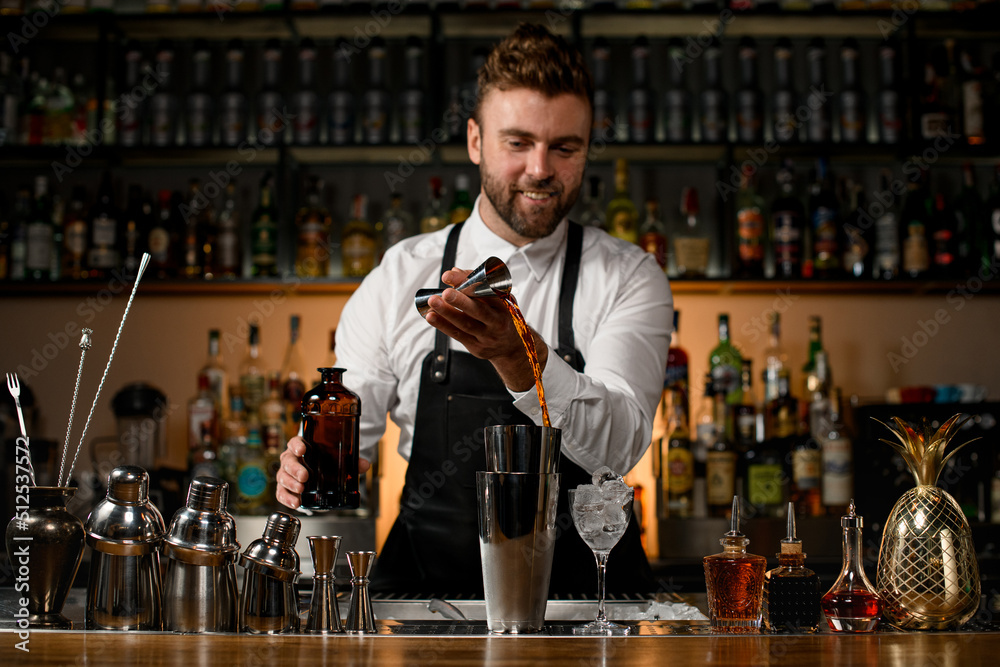 various shakers and bottles stand on the bar counter and male bartender accurate pours alcoholic drink into cup