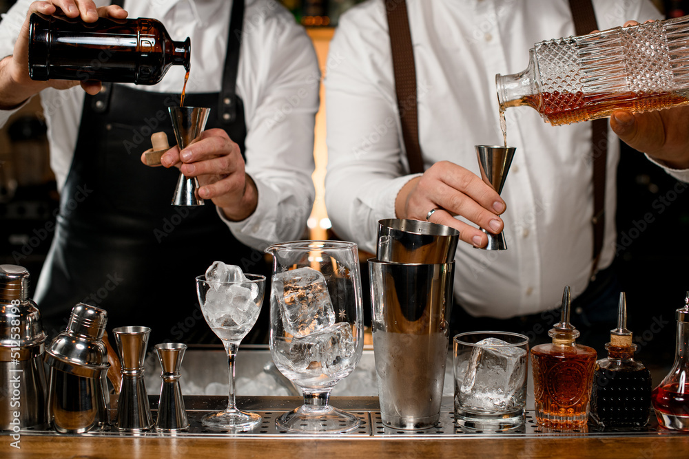 Close-up view of two male bartenders holding bottles and pours beverage into steel jiggers