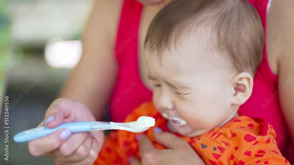 Mother feeds her child sitting in her arms. Baby eats white stuff and then closes eyes and frowns. Close up. Mother feeds her child sitting in her arms. Baby eats white stuff and then closes eyes and frowns. Close up.