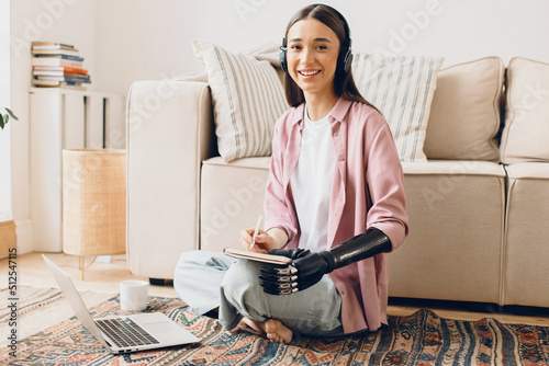 Smiling cheerful young female student listening to online English lesson in her wireless earphones, noting down in copybook, having bionic cyber hand, sitting against white sofa, in front of laptop photo