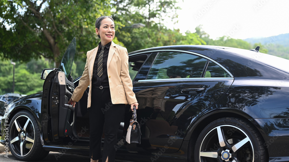 Attractive mature female entrepreneur holding bag and standing near her car at parking