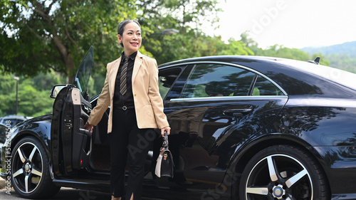 Attractive mature female entrepreneur holding bag and standing near her car at parking © Prathankarnpap