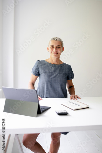 middle-aged businesswoman standing by white high table and working on tablet and wearing grey-blue dress and with short light hair