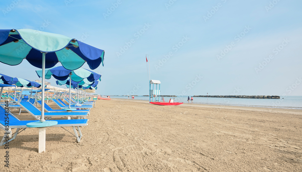 Fototapeta premium Deckchair with colorful umbrellas on a sunny summer day. In the background a lifeboat next to a lifeguard tower on the shore of the Adriatic Sea in Italy.