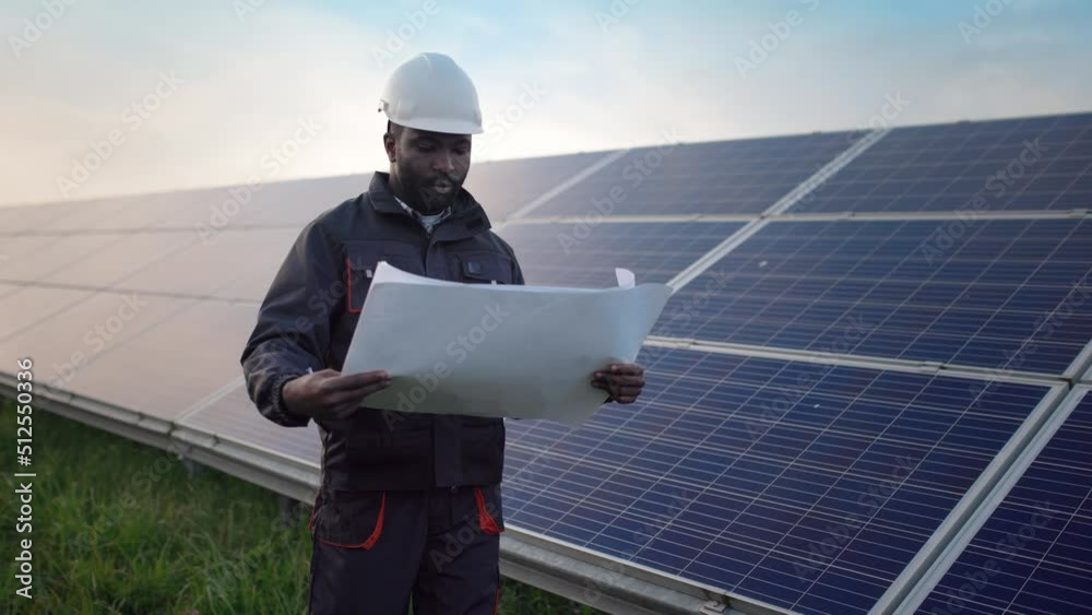 Young African-American engineer in protective helmet and uniform walks among the fields with solar panels with paper scheme in his hands studying location and serviceability of batteries. Young African-American engineer in protective helmet and uniform walks among the fields with solar panels with paper scheme in his hands studying location and serviceability of batteries.