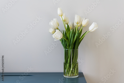 A bouquet of beautiful white tulips in a clear vase stands on a blue table. A light wall behind and a place for the text. Home interior