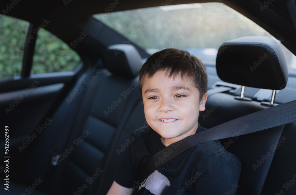 Happy School kid siting in safety car seat looking out with smiling face, Positive Child sitting in the back passenger seat with a safety belt, Young boy traveling to school by car.Back to school