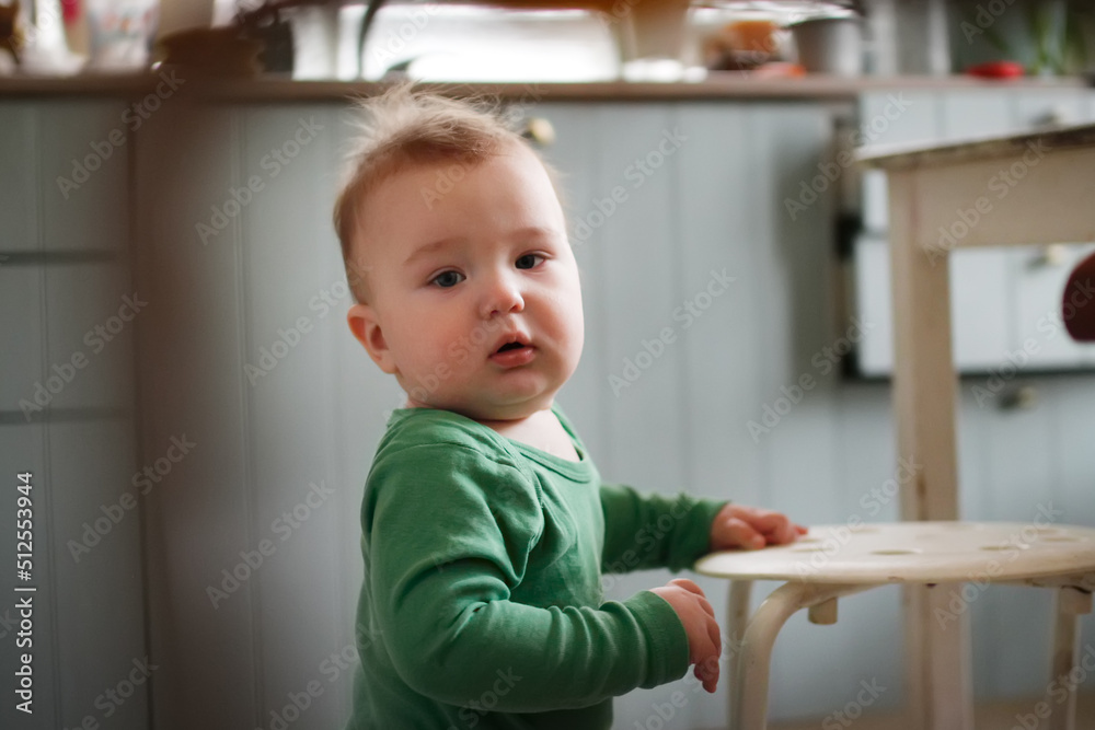Funny one-year-old toddler standing at stool in kitchen, real people in real interior Funny one-year-old toddler standing at stool in kitchen, real people in real interior