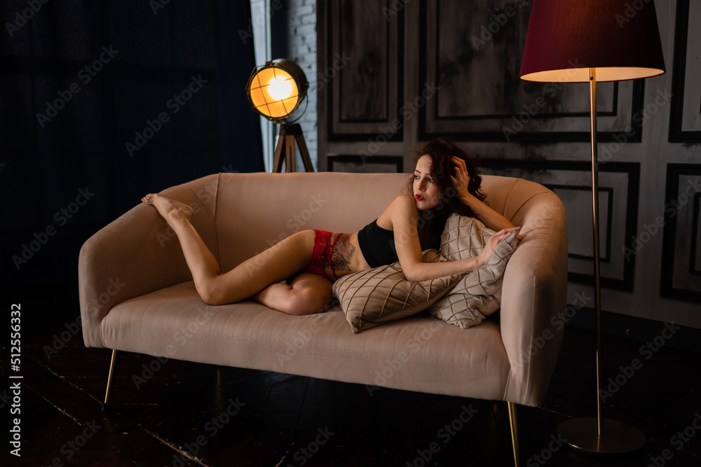Obraz premium incredibly sexy petite slender brunette with curly long hair in erotic transparent sexual lingerie underwear poses sensually lying on a sofa