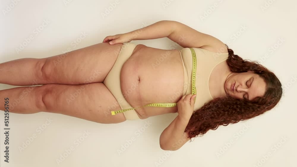 Vertical shot of long-haired curly overweight, adipose and fat woman in beige underwear, measuring the circumference of chest with yellow centimeter. Big size waist. Health care, obesity, overweight