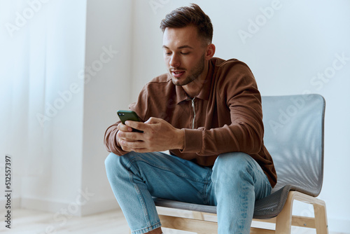 Friendly happy focused young man guy hold phone typing message chatting talking with family sitting on chair at home. Distance communication concept. Mobile App offer. Copy space