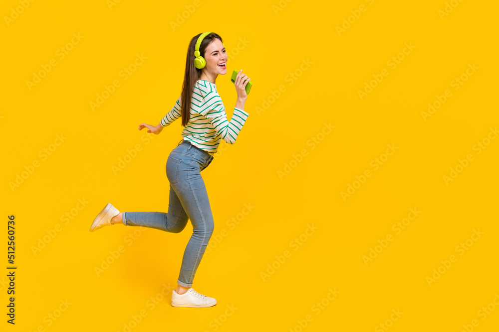 Full body profile side photo of laughing cheerful girl listen music in headset use phone as mic isolated on yellow color background Full body profile side photo of laughing cheerful girl listen music in headset use phone as mic isolated on yellow color background