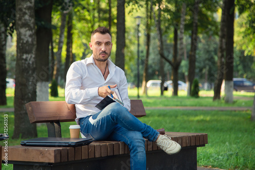 A serious man sits on a bench, makes plans, writes in a notebook. A young man on a background of green trees, a hot sunny summer day. Warm soft light, close-up.