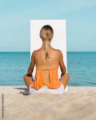 A blonde woman sits on a sandy beach in front of a white canvas.  The sea is in the background.