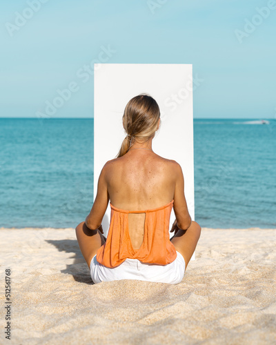 A blonde woman sits on a sandy beach in front of a white canvas.  The sea is in the background.