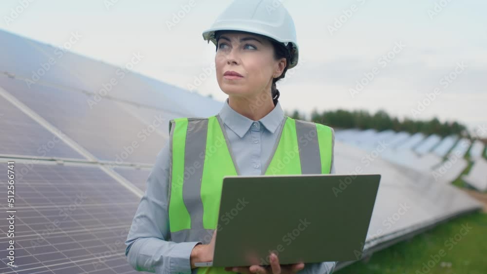 Beautiful caucasian female worker in signal vest and protective helmet stands near solar panels typing on laptop and looking around. Beautiful caucasian female worker in signal vest and protective helmet stands near solar panels typing on laptop and looking around.