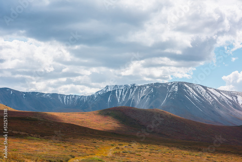 Motley autumn landscape with hills on high plateau and sunlit snowy mountain range under dramatic cloudy sky. Vivid autumn colors in mountains. Sunlight and shadows of clouds in changeable weather.