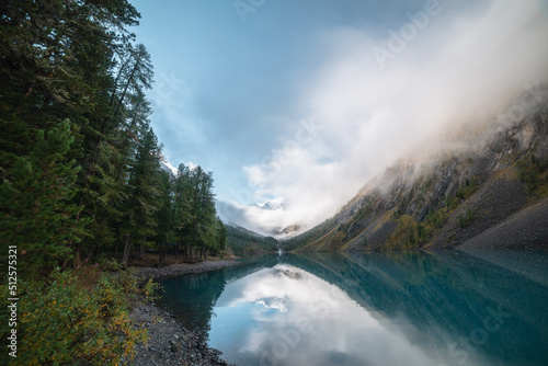 Scenic view to big pure mountain lake in autumn valley against snow mountain top in low clouds. Tranquil meditative landscape with snowy peak reflection in turquoise glacial lake in early morning.