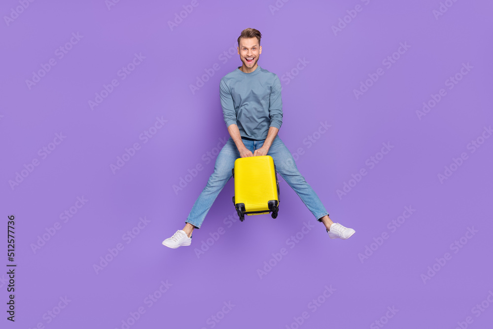 Photo of shiny excited guy dressed grey shirt jumping high holding luggage isolated purple color background Photo of shiny excited guy dressed grey shirt jumping high holding luggage isolated purple color background