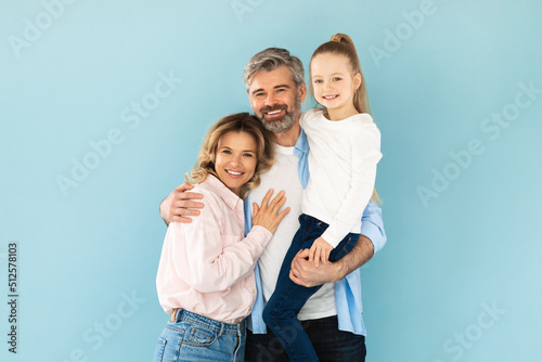 Man Holding Daughter In Arms Embracing Wife On Blue Background © Prostock-studio