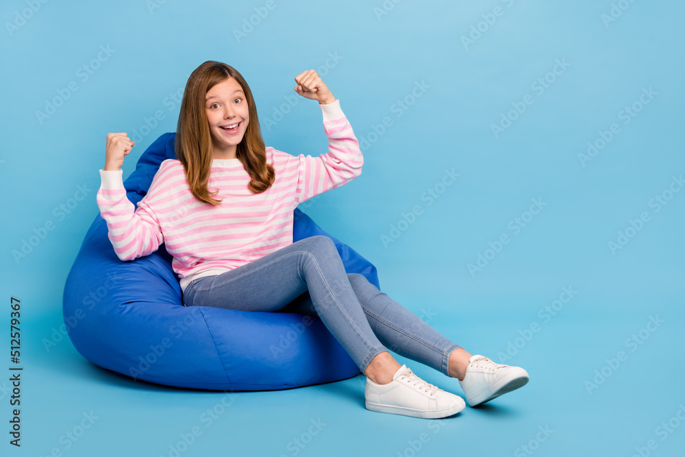 Full size portrait of delighted positive girl sit big soft chair raise fists success isolated on blue color background Full size portrait of delighted positive girl sit big soft chair raise fists success isolated on blue color background