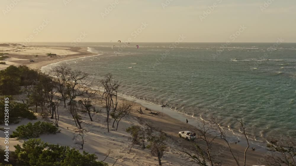 Offroad vehicles drive along beach toward kite surfers in sunset backdrop; drone Offroad vehicles drive along beach toward kite surfers in sunset backdrop; drone