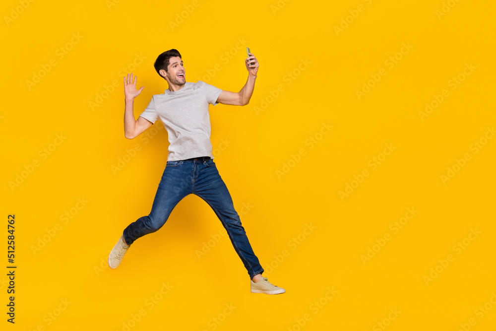 Full size photo of funny brunet millennial guy jump do selfie wear t-shirt jeans shoes isolated on yellow background