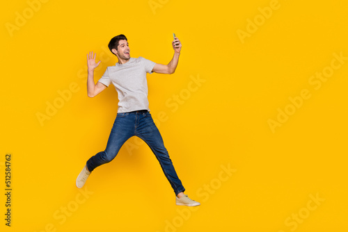 Full size photo of funny brunet millennial guy jump do selfie wear t-shirt jeans shoes isolated on yellow background © deagreez
