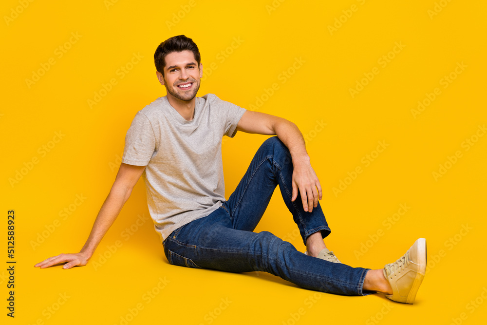 Full size photo of funky brunet young guy sit wear t-shirt jeans sneakers isolated on yellow background