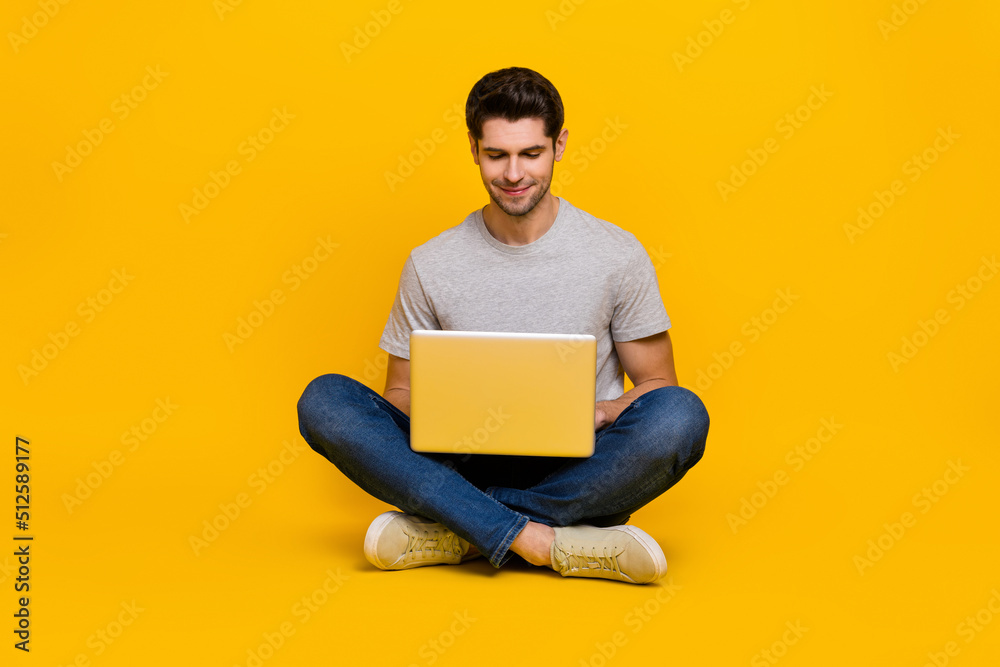 Full size photo of nice brunet young guy sit type laptop wear t-shirt jeans footwear isolated on yellow background